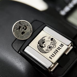 May include: A silver-colored, circular button with a crane design sits on a black camera. The camera's flash mount features a matching crane design and the word "FUJIFILM". The button and flash mount have a detailed, engraved appearance.