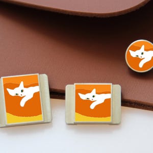 以下が含まれることがあります： 猫をテーマにしたアクセサリーセット。長方形のピン2つと丸いピン1つが含まれています。各ピンは、オレンジ色の背景に白い猫のデザインが特徴です。ピンは茶色の革の背景に配置されています。