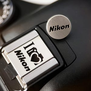 Könnte beinhalten: Nahaufnahme einer schwarzen Kamera mit silbernem Nikon-Logo und silbernem Zubehör mit dem Text "I ❤️ Nikon". Das Zifferblatt der Kamera ist im Hintergrund sichtbar. Das Bild ist gut beleuchtet und fokussiert.