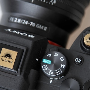 Puede incluir: Primer plano de una cámara Sony negra con una lente adjunta. La cámara presenta un dial de modo, botones y un logotipo Fujifilm dorado. La lente está etiquetada como "FE 28/24-70 GM".