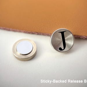 Op de afbeelding: Twee zilverkleurige, zelfklevende ontgrendelingsknoppen. E&eacute;n knop heeft een wit midden, de andere heeft een zwarte letter "J" op de voorkant. De knoppen liggen op een wit oppervlak, met een bruin, leerachtig materiaal op de achtergrond. Tekst: "Sticky-Backed Release Button".