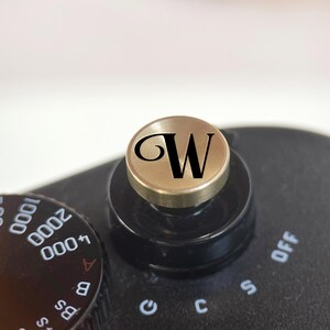 Op de afbeelding: Close-up van de zwarte en gouden bedieningselementen van een camera. Een goudkleurige wijzerplaat met de zwarte letter "W" staat bovenop een zwarte bedieningsknop. De instellingen van de camera zijn zichtbaar, inclusief cijfers en letters.