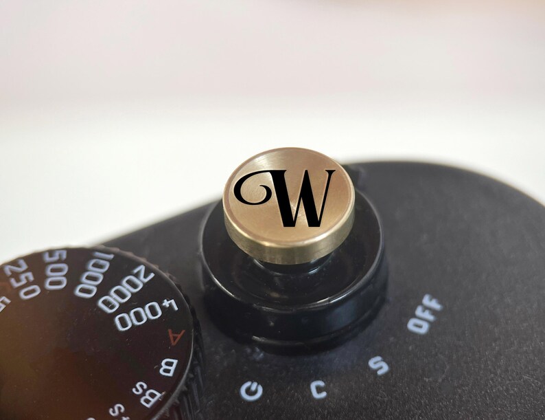 Op de afbeelding: Close-up van de zwarte en gouden bedieningselementen van een camera. Een goudkleurige wijzerplaat met de zwarte letter "W" staat bovenop een zwarte bedieningsknop. De instellingen van de camera zijn zichtbaar, inclusief cijfers en letters.