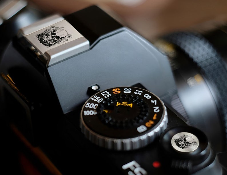 May include: A black and silver camera with a skull and floral design on the top. The camera has a dial with numbers from 12 to 64 and the word "ASA" on it.