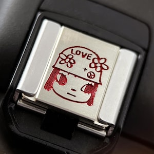 Puede incluir: Accesorio de metal plateado con una cara de dibujos animados roja y la palabra "LOVE" encima. La cara tiene un casco con flores y un símbolo de la paz. El accesorio está sujeto a un dispositivo negro.