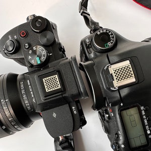 Könnte beinhalten: Zwei schwarze Digitalkameras mit Objektiven sind in entgegengesetzten Winkeln positioniert. Eine Kamera ist eine Sony α7 mit einem 24-70mm-Objektiv. Die andere Kamera hat ein Canon-Objektiv. Beide Kameras haben verschiedene Knöpfe und Räder.