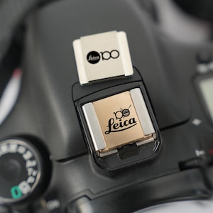 May include: Close-up of a black camera body with a silver and gold flash shoe cover. The cover has the Leica logo and the number 100 printed on it. The camera's dial and other controls are visible.