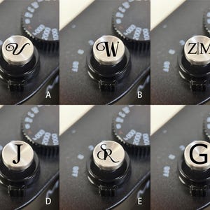 Op de afbeelding: Close-up van een zwarte camerabody met een zilveren draaiknop met de letter "V". De draaiknop is omgeven door een zwarte bedieningsring met witte markeringen. De achtergrond toont andere draaiknoppen met cijfers.