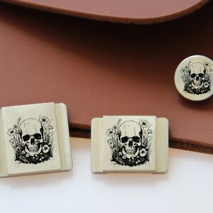 May include: Three silver metal skull and flower designs. The smallest design is round and the two larger designs are square.