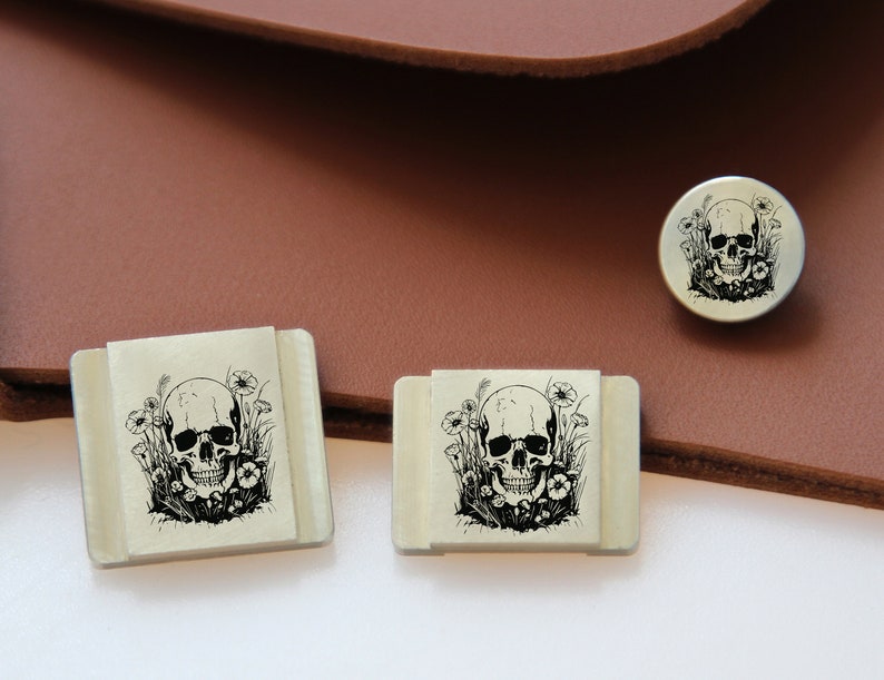 May include: Three silver metal skull and flower designs. The smallest design is round and the two larger designs are square.