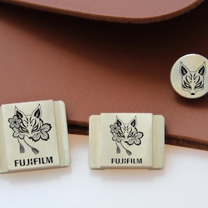 Puede incluir: Una colección de artículos decorativos con un diseño estilizado de cara de zorro. Los artículos incluyen dos piezas rectangulares y un botón redondo, todos con la palabra "FUJIFILM" impresa. El diseño del zorro es en blanco y negro.
