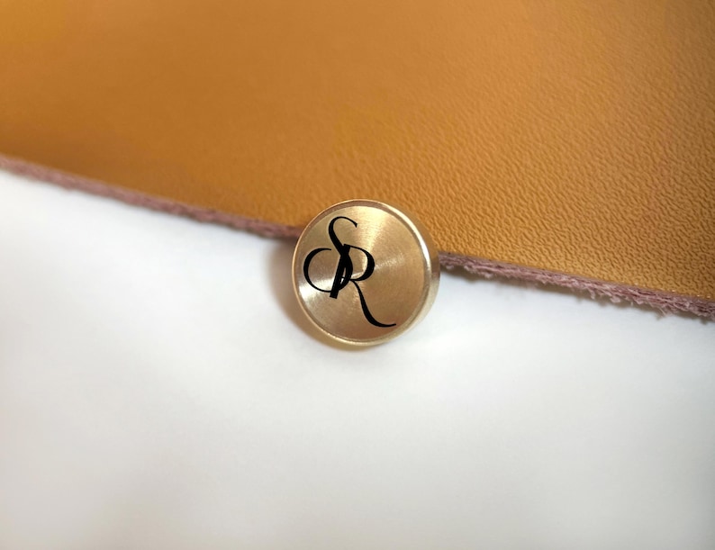 Op de afbeelding: Een kleine, ronde, goudkleurige knoop met de zwarte letters "SR" in een cursief lettertype. De knoop staat tegen een achtergrond van een getextureerd, bruin oppervlak en een wit oppervlak.