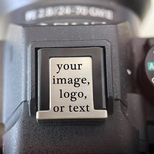 May include: A silver-toned metal hot shoe cover for a camera, featuring a rectangular plate with the engraved black text 'your image, logo, or text' centered on the surface. The cover is securely inserted into the black camera hot shoe mount.