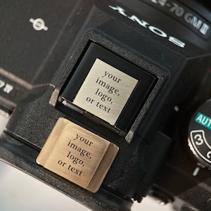 Könnte beinhalten: Nahaufnahme einer schwarzen Kamera mit silbernem Objektiv. Die Kamera hat ein Einstellrad mit Einstellungen und den Text "AUTO". Zwei kleine rechteckige Platten sind angebracht, jede mit dem Text "your image, logo, or text".