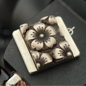 May include: A silver-toned metal charm with a square shape and a detailed design of five cherry blossoms.