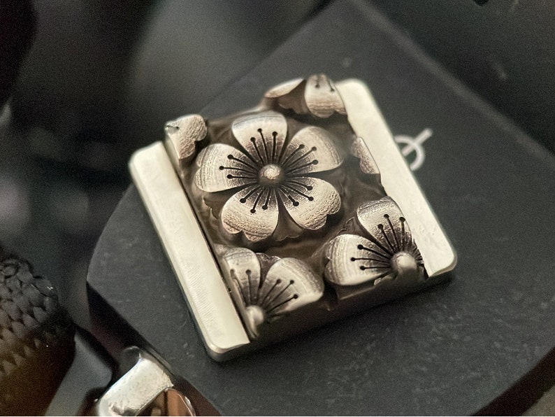 May include: A silver-toned metal charm with a square shape and a detailed design of five cherry blossoms.