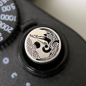 Puede incluir: Primer plano de una cámara negra con un botón plateado con un diseño detallado de dragón. El botón es circular y se encuentra en un dial negro. La configuración de la cámara es visible en el fondo.