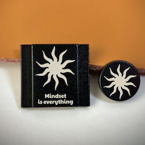 Op de afbeelding: Een zwarte vierkante speld en een zwarte ronde speld, beide voorzien van een witte zongrafiek. De vierkante speld bevat de witte tekst "Mindset is everything" onder de zon. Beide items hebben een matte afwerking en een strak ontwerp.