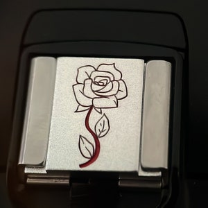 May include: A silver-colored square tile with a red and white rose design. The rose is outlined in black and has a red stem and leaves.