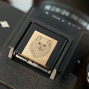 May include: A black camera with a silver metal plate featuring a line drawing of a cat's face. The plate is attached to the top of the camera.