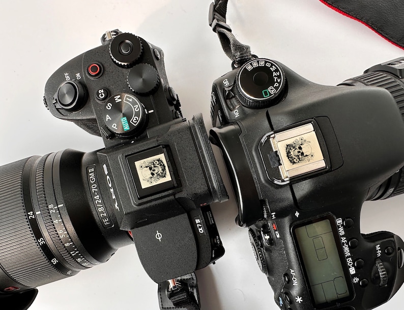 May include: A black Sony Alpha camera with a 24-70mm lens attached. The camera has a skull sticker on the top. A black Canon camera is also in the image, with a skull sticker on the top.