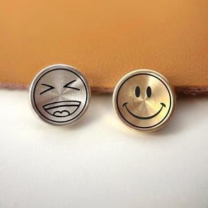 May include: Two round metal pins with smiley faces. One pin is silver with a laughing face design, and the other is gold with a smiling face design. The pins are small and circular, with black outlines and details.