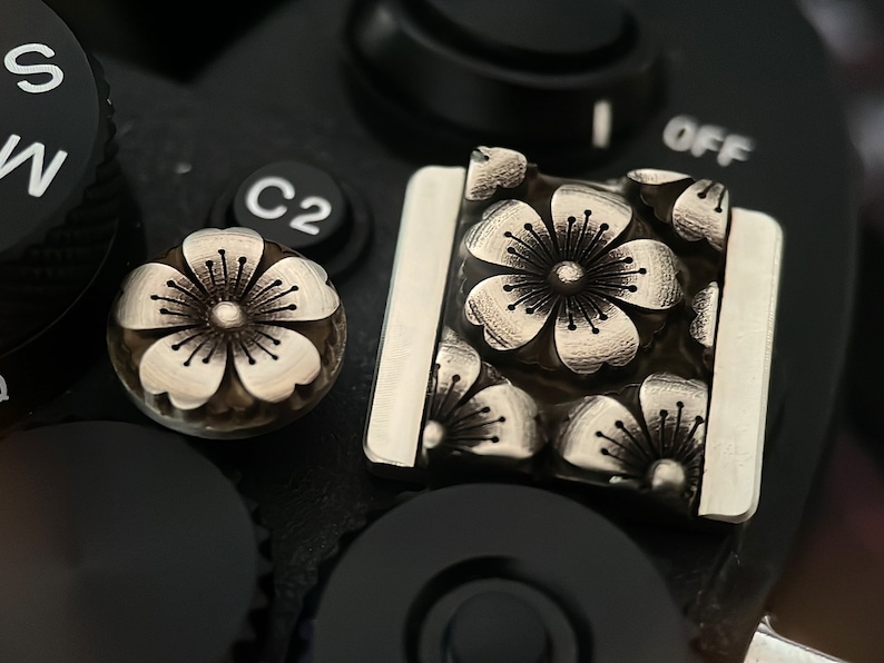May include: A close-up of a silver camera accessory with a floral design. The accessory has a rectangular shape and is made of metal. The flowers are embossed and have a detailed design.