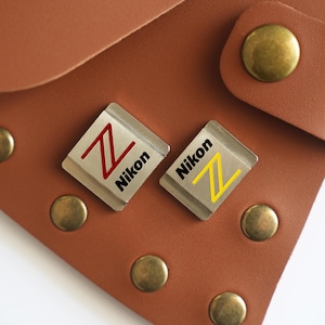 Puede incluir: Dos pines plateados de Nikon con un diseño "Z" rojo y amarillo, sobre un fondo de cuero marrón con remaches de latón. Los pines tienen el logotipo de Nikon en negro.