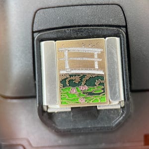 May include: A rectangular metal charm featuring a silver-toned torii gate design positioned above a green pond with pink water lilies. The charm has a metallic finish and is designed to be attached to a bracelet or similar jewelry piece.