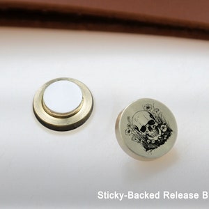 May include: A round, silver-colored button with a black and white skull and flower design. The button has a sticky back and is labeled "Sticky-Backed Release Button".