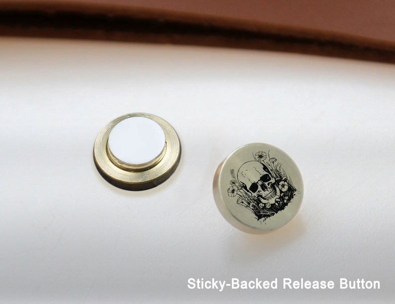 May include: A round, silver-colored button with a black and white skull and flower design. The button has a sticky back and is labeled "Sticky-Backed Release Button".