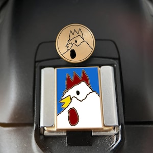 May include: A circular brass-toned metal pin featuring an etched chicken head design sits above a rectangular enamel pin. The rectangular pin displays a white chicken with a red comb and wattle, a yellow beak, and a black eye against a blue background.