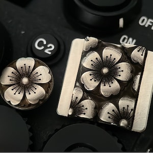 May include: A silver camera button with a floral design. The button is shaped like a flower with five petals and a center circle. The button is surrounded by a black background.