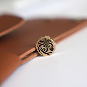May include: A gold-colored metal disc with a black and white swirling pattern. The disc is sitting on a brown leather surface.