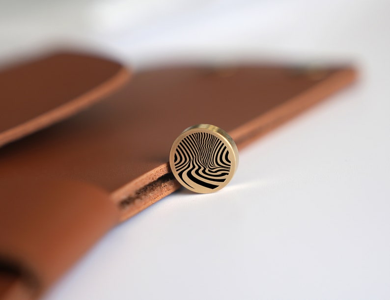 May include: A gold-colored metal disc with a black and white swirling pattern. The disc is sitting on a brown leather surface.
