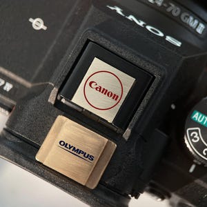 Puede incluir: Primer plano de un cuerpo de cámara negro con el logotipo de Canon en un círculo rojo. Un logotipo de Olympus es visible debajo. La cámara tiene un dial con ajustes, incluido AUTO. La lente es una 24-70 GM II.