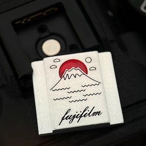 May include: A silver camera strap with a red and white illustration of Mount Fuji and the words "fujifilm" in black.