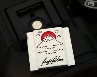 May include: A silver camera strap with a red and white illustration of Mount Fuji and the words "fujifilm" in black.