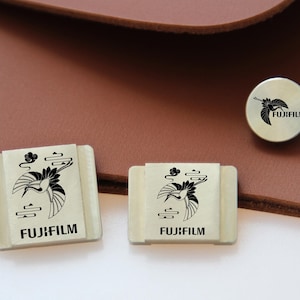 Puede incluir: Accesorios Fujifilm rectangulares y circulares de color crema con un diseño de grulla negra. La grulla está representada en vuelo con nubes estilizadas. Los accesorios están colocados sobre un fondo de cuero marrón.