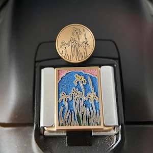 Peut inclure: Une épingle ronde dorée avec un motif de fleurs d'iris. En dessous, une pièce rectangulaire en émail coloré représente des iris sur fond de ciel bleu. L'épingle et la pièce en émail sont posées sur une surface noire.