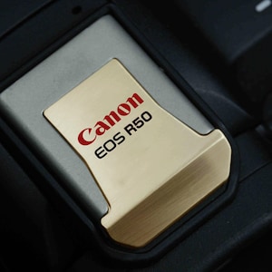May include: A close-up of a Canon EOS R50 camera memory card slot cover. The cover is gold-coloured with the Canon logo and EOS R50 model name in red lettering. The surrounding area is black and grey.