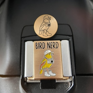 May include: A rectangular camera hot shoe cover in brushed gold metal featuring a yellow bird wearing a white hat and boots while carrying a camera, with the text BIRD NERD printed above. A matching circular gold metal pin is placed above it.