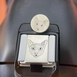 Puede incluir: Una tapa de zapata para cámara de metal plateado con un grabado de líneas detallado de una cara de gato. El accesorio está colocado en la zapata de una cámara negra, con una pieza metálica circular a juego situada justo encima.