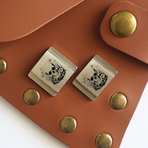 May include: Two silver metal keychains with a skull and floral design. The keychains are on a brown leather surface.