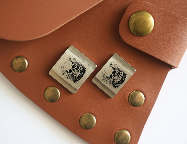 May include: Two silver metal keychains with a skull and floral design. The keychains are on a brown leather surface.