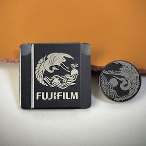 Op de afbeelding: Twee zwarte Fujifilm-accessoires met zilveren ontwerpen. Eén is rechthoekig met het Fujifilm-logo en een kraanvogel over golven. De andere is een ronde pin met een kraanvogel- en zonontwerp.
