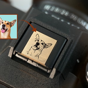 May include: A black camera with a small, engraved metal plate featuring a black and white image of a dog. The plate is attached to the top of the camera, near the lens.