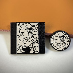 Könnte beinhalten: Zwei Objekte mit einem schwarz-weißen Grafikdesign einer Figur mit Stab auf cremefarbenem Hintergrund. Ein Objekt ist quadratisch, das andere rund. Möglicherweise Schmuck oder Dekorationsartikel.