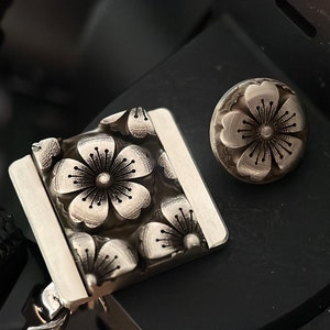 May include: A silver camera strap with a floral design. The strap has a square metal piece with a raised design of four flowers. The metal piece is attached to a black strap. There is a separate silver flower-shaped button.