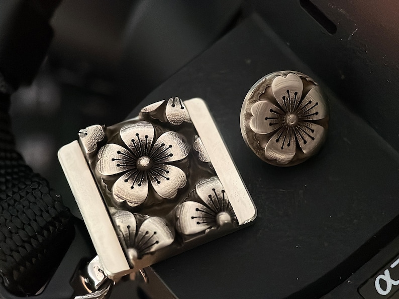 May include: A silver camera strap with a floral design. The strap has a square metal piece with a raised design of four flowers. The metal piece is attached to a black strap. There is a separate silver flower-shaped button.
